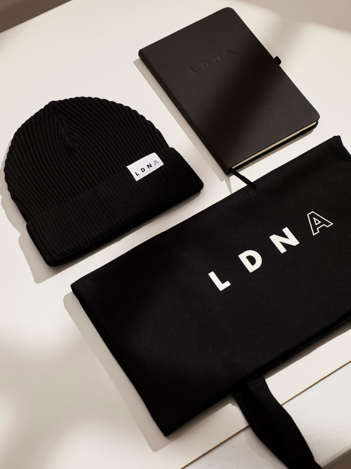 LDNA black fisherman cuffed beanie, LDNA black tote bag and LDNA black hardcover notebook neatly arranged on a white table.