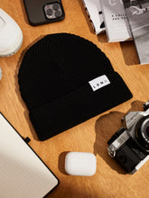 Load image into Gallery viewer,  LDNA London black fisherman style beanie with Canon camera, iPhone, magazines and LDNA lined paper notebook on wooden table.
