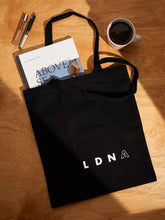 Load image into Gallery viewer, LDNA London black tote bag on a wooden table with lifestyle items such as magazine, coffee,  sunglasses and pen.
